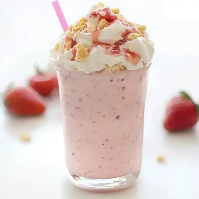 Ein rosa Milchshake garniert mit Schlagsahne, zerkleinerten Keksen und Erdbeersirup, serviert mit einem Strohhalm, daneben frische Erdbeeren.