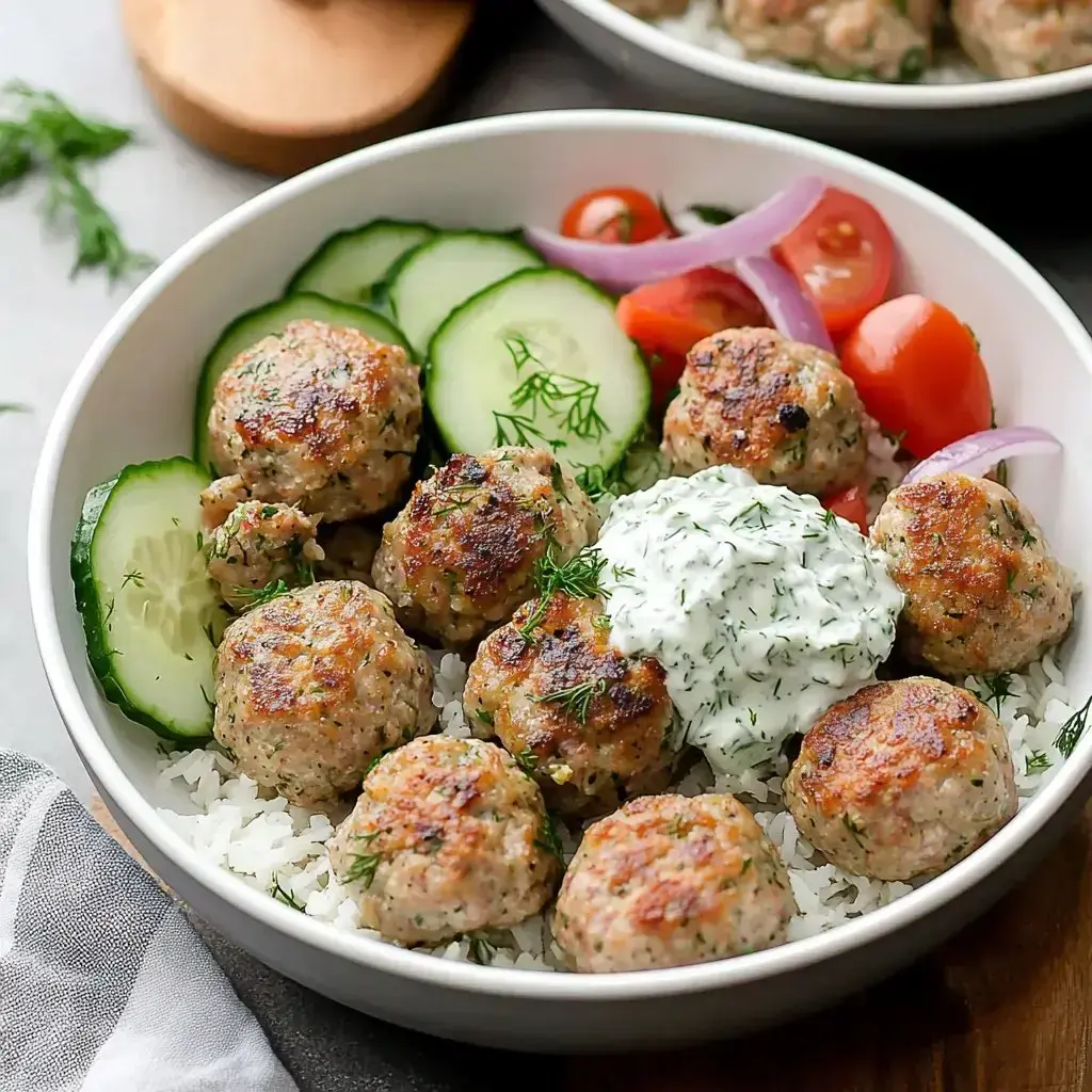 Eine Schüssel mit Reis, belegt mit goldbraunen Hackbällchen, geschnittenen Gurken, Kirschtomaten, roten Zwiebeln und einem Klecks cremiger Kräutersauce.