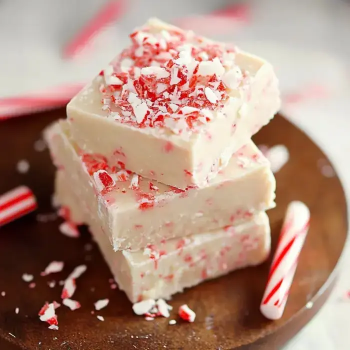 Ein Stapel cremiger weißer Pfefferminz-Fudge mit zerkleinerten Zuckerstangen bestreut, daneben kleine Zuckerstangen auf einer Holzplatte.