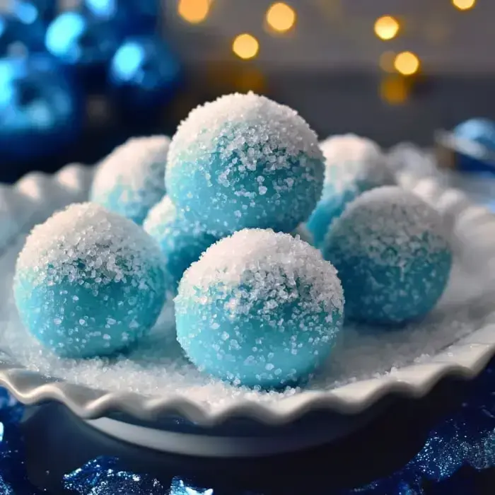 Ein Teller mit blauen, zuckerbestäubten Kugeln vor einem festlichen Hintergrund mit blauen Christbaumkugeln und weicher Beleuchtung.