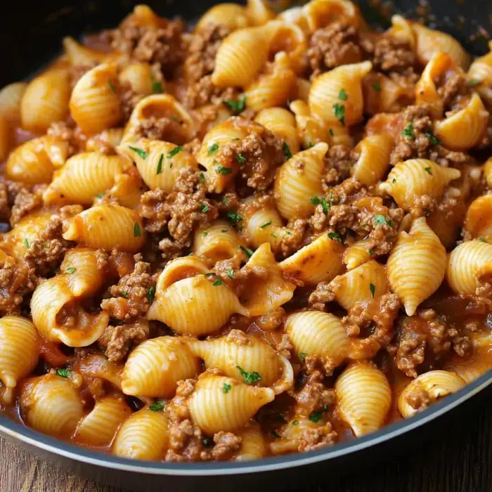 Eine Pfanne gefüllt mit gekochten Muschelnudeln, vermischt mit Hackfleisch und einer reichhaltigen Tomatensauce, garniert mit Petersilie.