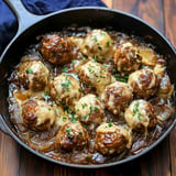 Eine Gusseisenpfanne gefüllt mit Hackbällchen, überbacken mit geschmolzenem Käse und bestreut mit frischer Petersilie, in einer würzigen braunen Sauce.