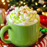 Eine leuchtend grüne Tasse gefüllt mit Schlagsahne, Mini-Marshmallows und grünem Sirup vor weihnachtlichem Hintergrund mit Dekoration.