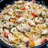 Eine Nahaufnahme eines cremigen Hähnchen-Pasta-Gerichts mit Farfalle-Nudeln, gewürfeltem Gemüse und frischer Petersilie garniert.