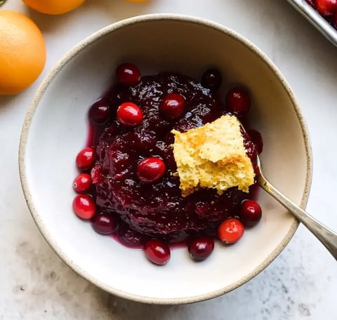 Eine Schüssel Cranberry-Sauce garniert mit frischen Cranberries und einem Stück Maisbrot, begleitet von ganzen Orangen im Hintergrund.